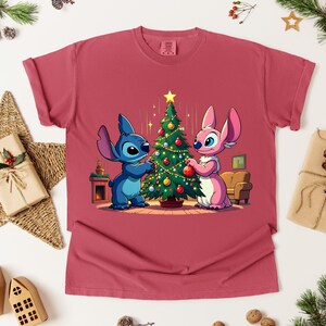 May include: A maroon t-shirt featuring a cartoon illustration of Stitch and Angel decorating a Christmas tree. The design includes a star, ornaments, and a cozy interior scene. The shirt is displayed on a white surface with holiday decorations.