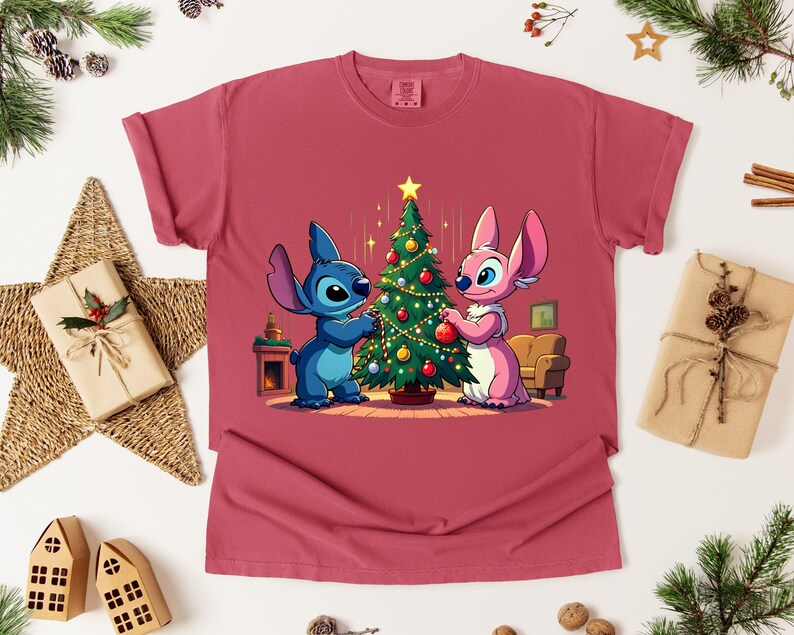 May include: A maroon t-shirt featuring a cartoon illustration of Stitch and Angel decorating a Christmas tree. The design includes a star, ornaments, and a cozy interior scene. The shirt is displayed on a white surface with holiday decorations.