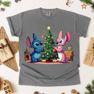 May include: Gray t-shirt featuring a cartoon illustration of Stitch and Angel decorating a Christmas tree. The characters are in blue and pink, with a decorated tree and a cozy interior scene. The shirt is a comfortable, casual style.