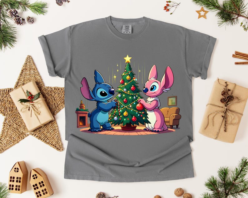 May include: Gray t-shirt featuring a cartoon illustration of Stitch and Angel decorating a Christmas tree. The characters are in blue and pink, with a decorated tree and a cozy interior scene. The shirt is a comfortable, casual style.
