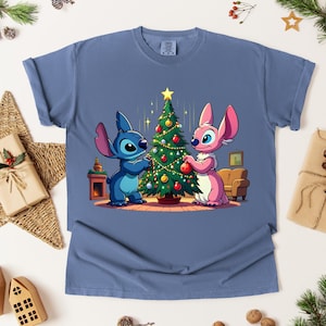 May include: A blue t-shirt featuring a cartoon illustration of two characters decorating a Christmas tree. One character is blue, the other is pink. The tree is decorated with ornaments and a star.