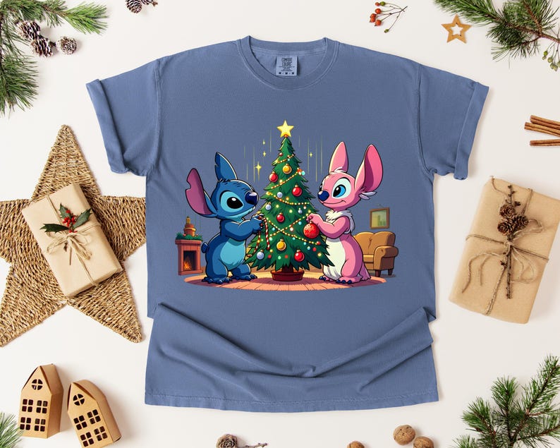 May include: A blue t-shirt featuring a cartoon illustration of two characters decorating a Christmas tree. One character is blue, the other is pink. The tree is decorated with ornaments and a star.
