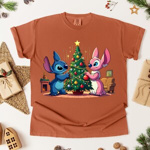 May include: Rust-colored t-shirt featuring a cartoon illustration of two characters decorating a Christmas tree. One character is blue, the other is pink. The tree is adorned with ornaments and a star.