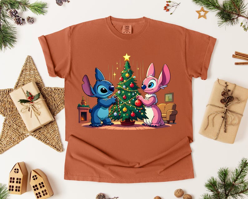 May include: Rust-colored t-shirt featuring a cartoon illustration of two characters decorating a Christmas tree. One character is blue, the other is pink. The tree is adorned with ornaments and a star.