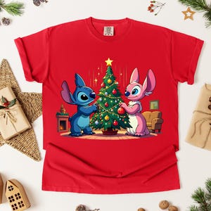 May include: Red t-shirt featuring a cartoon image of Stitch and Angel decorating a Christmas tree. The characters are blue and pink, and the tree is adorned with ornaments. The shirt is surrounded by Christmas decorations.