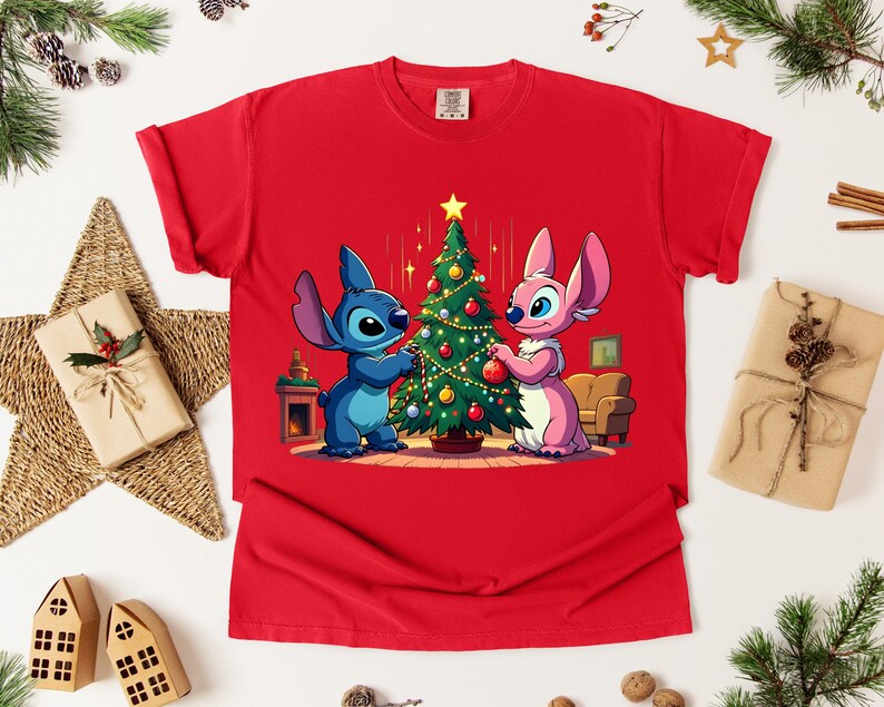 May include: Red t-shirt featuring a cartoon image of Stitch and Angel decorating a Christmas tree. The characters are blue and pink, and the tree is adorned with ornaments. The shirt is surrounded by Christmas decorations.