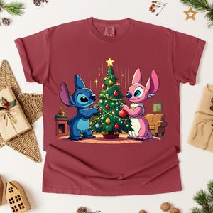 May include: A maroon t-shirt featuring a cartoon illustration of two characters decorating a Christmas tree. One character is blue, the other is pink. The tree is decorated with ornaments and a star.