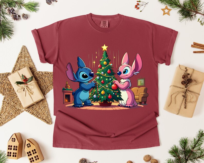 May include: A maroon t-shirt featuring a cartoon illustration of two characters decorating a Christmas tree. One character is blue, the other is pink. The tree is decorated with ornaments and a star.