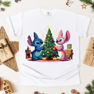 May include: White t-shirt featuring a cartoon illustration of two characters decorating a Christmas tree. One character is blue, the other is pink. The tree is decorated with ornaments and a star. The shirt is surrounded by Christmas decorations.