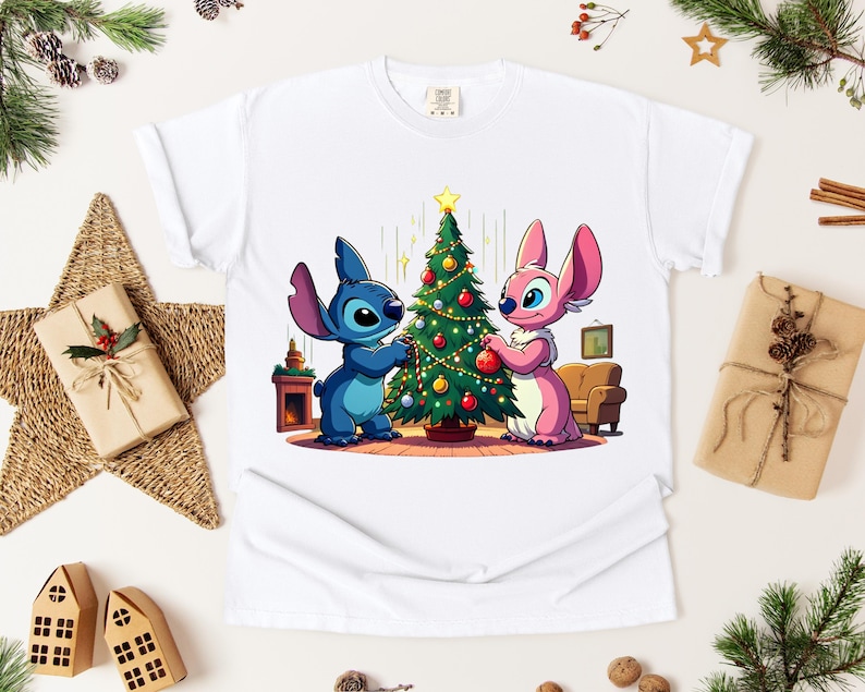 May include: White t-shirt featuring a cartoon illustration of two characters decorating a Christmas tree. One character is blue, the other is pink. The tree is decorated with ornaments and a star. The shirt is surrounded by Christmas decorations.