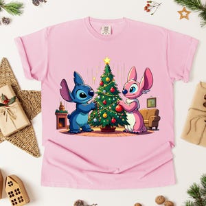 May include: Pink t-shirt featuring a cartoon illustration of Stitch and Angel decorating a Christmas tree. The characters are blue and pink, with the tree adorned with ornaments. A festive holiday design.