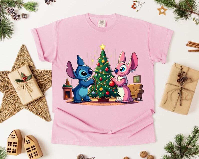 May include: Pink t-shirt featuring a cartoon illustration of Stitch and Angel decorating a Christmas tree. The characters are blue and pink, with the tree adorned with ornaments. A festive holiday design.