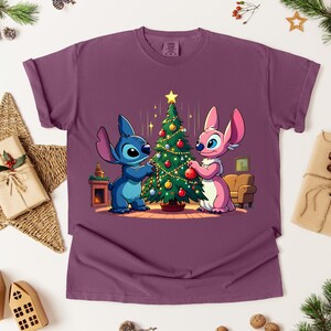 May include: A maroon t-shirt featuring a cartoon illustration of two characters decorating a Christmas tree. One character is blue, the other is pink. The tree is decorated with ornaments and a star. Festive holiday apparel.