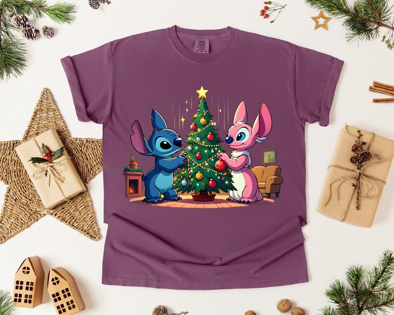 May include: A maroon t-shirt featuring a cartoon illustration of two characters decorating a Christmas tree. One character is blue, the other is pink. The tree is decorated with ornaments and a star. Festive holiday apparel.