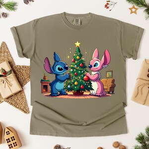 May include: Olive green t-shirt featuring a cartoon illustration of a blue and pink alien decorating a Christmas tree. The design includes a star, ornaments, and a cozy living room scene. Festive and holiday-themed.