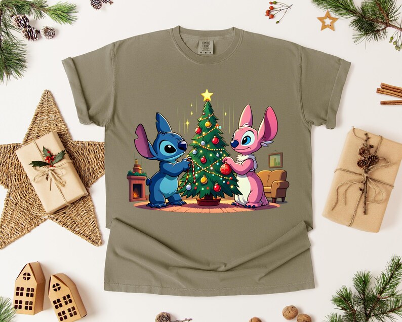 May include: Olive green t-shirt featuring a cartoon illustration of a blue and pink alien decorating a Christmas tree. The design includes a star, ornaments, and a cozy living room scene. Festive and holiday-themed.