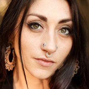 May include: Portrait of a person with dark hair and green eyes, wearing multiple pieces of facial jewelry. The jewelry includes a septum ring and nose studs. The person is also wearing large, ornate earrings. The person is wearing a black top.