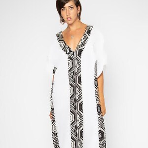 Peut inclure: Longue robe blanche avec un col en V et des manches courtes. La robe pr&eacute;sente un motif g&eacute;om&eacute;trique noir et blanc le long de l'encolure et sur le devant. La robe est longue et faite d'un tissu l&eacute;ger.