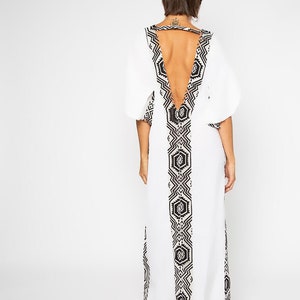 Peut inclure: Longue robe blanche avec un dos nu et un motif g&eacute;om&eacute;trique noir et blanc. La robe a des manches courtes et bouffantes et un motif d&eacute;coratif dans le dos et sur le devant. La robe est longue.