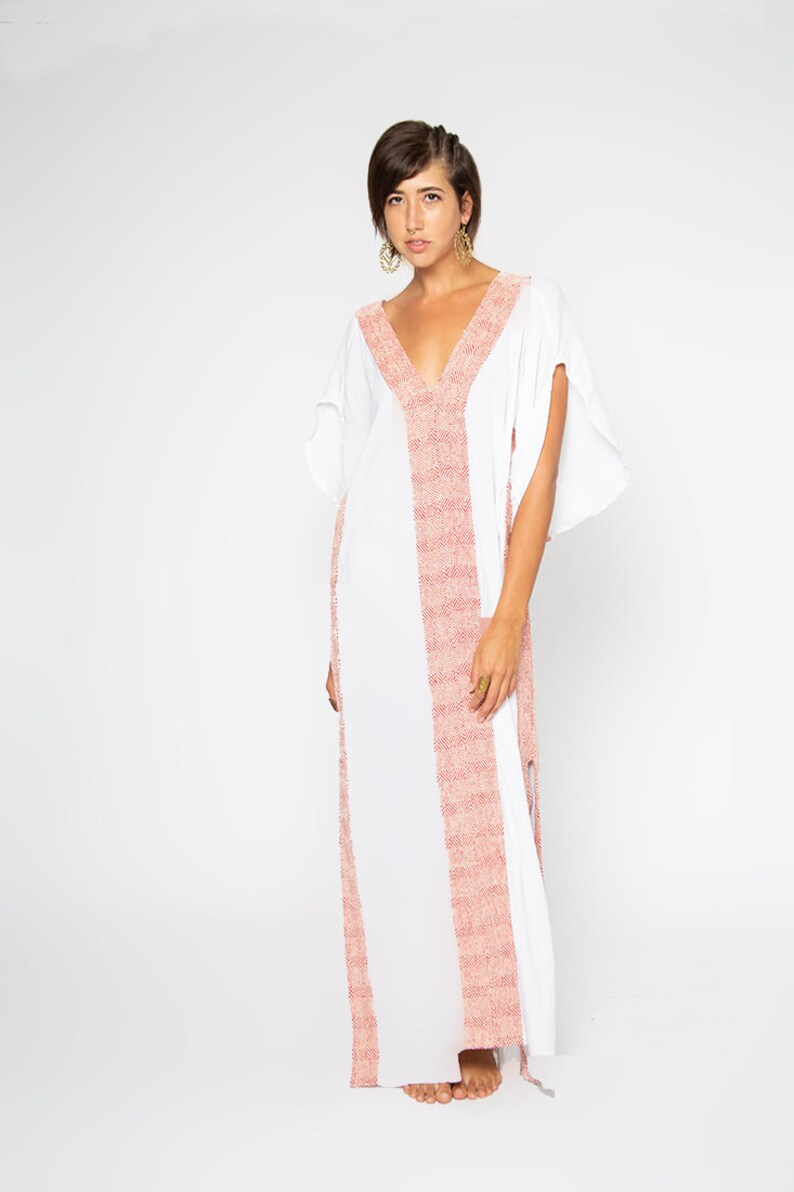 Peut inclure: Une robe caftan blanche avec un col en V profond et des manches courtes. La robe pr&eacute;sente une bande verticale de tissu rose tiss&eacute; au centre et le long de l'encolure. La mod&egrave;le porte des boucles d'oreilles et des bagues dor&eacute;es.