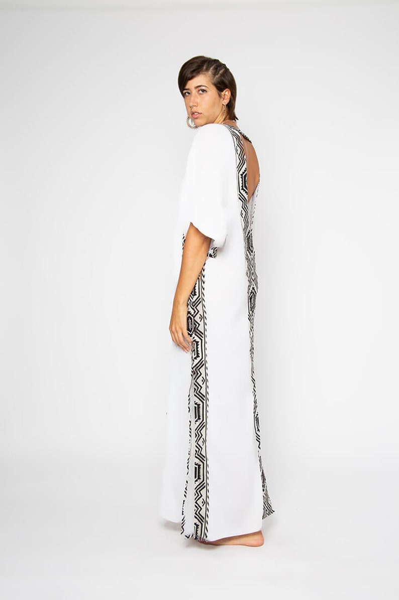 Peut inclure: Une robe longue blanche orn&eacute;e d'un motif g&eacute;om&eacute;trique noir et blanc sur les c&ocirc;t&eacute;s et le dos. La robe a des manches courtes et un dos nu. Le mod&egrave;le est tourn&eacute; dos &agrave; la cam&eacute;ra.
