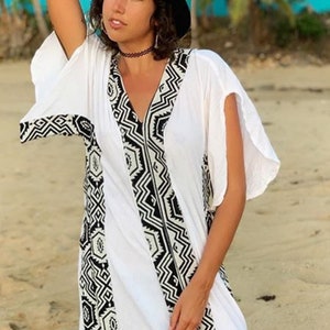 Peut inclure: Longue robe blanche &agrave; col en V et manches courtes. La robe pr&eacute;sente un motif g&eacute;om&eacute;trique noir et blanc sur les bords et une fente sur le c&ocirc;t&eacute;. Un chapeau noir et un ras du cou sont &eacute;galement visibles.