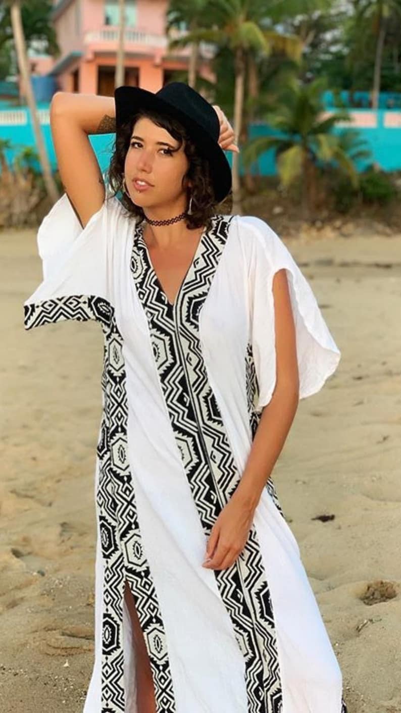 Peut inclure: Longue robe blanche &agrave; col en V et manches courtes. La robe pr&eacute;sente un motif g&eacute;om&eacute;trique noir et blanc sur les bords et une fente sur le c&ocirc;t&eacute;. Un chapeau noir et un ras du cou sont &eacute;galement visibles.