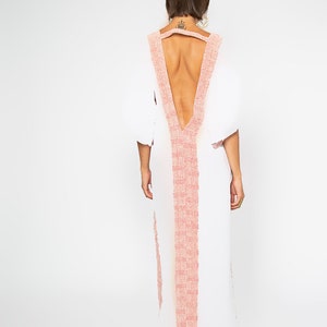 Peut inclure: Une robe blanche longue avec un d&eacute;collet&eacute; en V plongeant et un dos nu. La robe pr&eacute;sente une bordure rose clair textur&eacute;e le long de l'encolure et au centre. Les manches sont bouffantes. La robe est port&eacute;e par une personne.