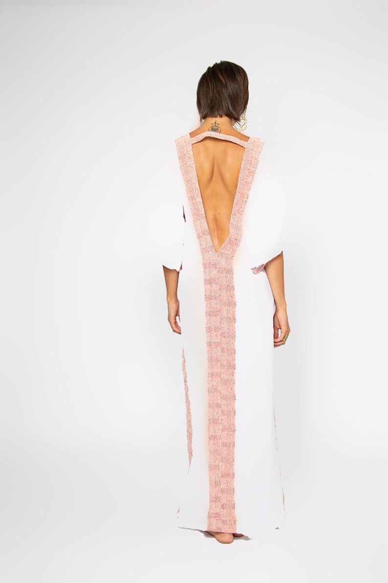 Peut inclure: Une robe blanche longue avec un d&eacute;collet&eacute; en V plongeant et un dos nu. La robe pr&eacute;sente une bordure rose clair textur&eacute;e le long de l'encolure et au centre. Les manches sont bouffantes. La robe est port&eacute;e par une personne.