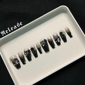 Puede incluir: Un conjunto de diez uñas postizas en forma de ataúd. Cada uña presenta un diseño degradado que va de transparente a negro, adornado con rosas negras y detalles brillantes. La palabra "Release" es visible en la imagen.