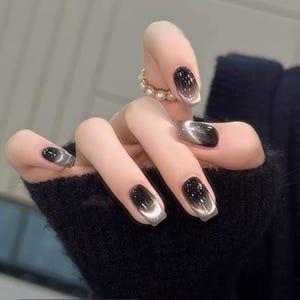 May include: A hand with long, square-shaped nails. The nails feature a gradient design, transitioning from black to silver. A gold ring with pearls adorns a finger. The nails are a stylish accessory.