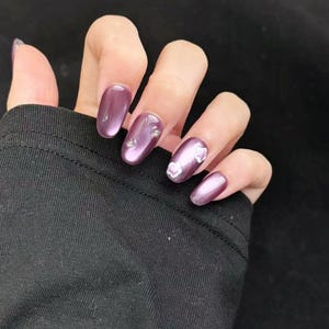 Puede incluir: Uñas ovaladas con esmalte morado con acabado nacarado. Algunas uñas presentan pequeños adornos transparentes y detalles de flores blancas. Las uñas están en una mano, con una manga negra de fondo.