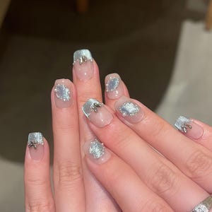May include: Close-up of a hand with manicured nails. The nails are painted with a clear base and feature silver glitter accents. Some nails have small silver embellishments. A silver ring is visible on the ring finger.