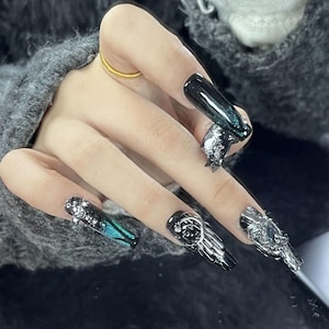 Può includere: Unghie finte lunghe, nere, a forma di bara con decorazioni argentate. Le unghie presentano ali metalliche, accenti floreali e gemme scintillanti. Alcune unghie hanno una sfumatura di verde acqua e nero.