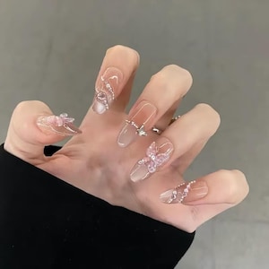 May include: A hand showing long, clear acrylic nails. Each nail is decorated with intricate designs, including silver embellishments, pink butterflies, and rhinestones. The nails have a natural, gradient colour scheme.