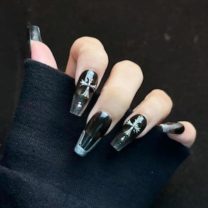 May include: Long, coffin-shaped acrylic nails with a black and silver gradient design. Some nails feature a silver cross charm. The hand is wearing a black sleeve.
