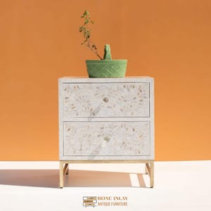 May include: A white two-drawer nightstand with floral inlay and gold-colored legs. A green ceramic planter with a small plant sits on top. The nightstand has crystal knobs and the text "BONE INLAY ANTIQUE FURNITURE" at the bottom.