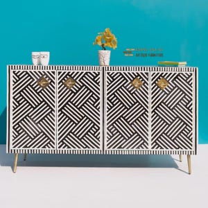 May include: A black and white geometric patterned cabinet with gold-colored hardware and legs. The cabinet has four doors and is topped with decorative items, including a small plant and two white mugs. The background is a solid teal color.
