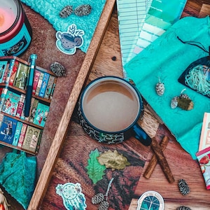 Op de afbeelding: Een houten dienblad met een kop koffie, een kaars en een notitieblok met boeken als thema. Bevat ook theezakjes, decoratieve items en een kleine cadeauzak. De items zijn gerangschikt op een houten oppervlak.