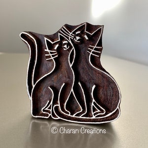 May include: A wooden block carving of two cats sitting side-by-side. The cats are outlined in white and the block is a dark brown color.