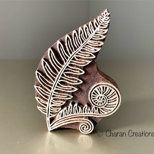 Puede incluir: Un bloque de madera marrón con un diseño tallado de hoja de helecho. El diseño incluye una hoja detallada y una espiral. Los detalles blancos contrastan con la madera. Para impresión o artesanía.