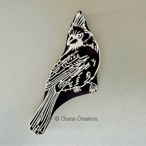 May include: A black and white wooden block carving of a bird with a long tail. The bird is facing to the left and has a crest on its head. The carving is on a light grey background. © Charan Creations