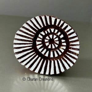 May include: A circular wooden printing block featuring a carved spiral pattern with white paint on the raised surfaces. The design consists of radiating lines that form a concentric circular motif. The text Charan Creations is printed below.