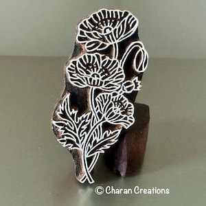 Può includere: Un blocco per stampa in legno marrone scuro con un disegno floreale intagliato di tre papaveri con steli e foglie. Le linee bianche in rilievo definiscono l'intricato motivo botanico. Il testo Charan Creations è stampato nella parte inferiore.