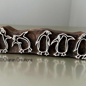 May include: A wooden block with five penguin designs carved into it. The penguins are outlined in white against the dark brown wood. The text "© Charan Creations" is printed on the block.