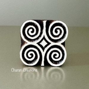 May include: A square wooden printing block featuring a symmetrical design of four white spiral patterns against a dark brown background. The text Charan Creations is printed in white in the lower left corner of the object's front face.