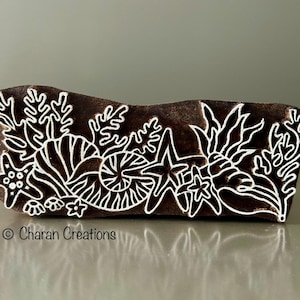 May include: A wooden block printing stamp with a white design of seashells, starfish, and seaweed on a brown background. The stamp is made for creating patterns on fabric.