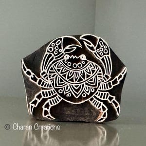 Könnte beinhalten: Ein dunkler Holzblock mit einem weißen Krebsdesign. Der Krebs ist aufwendig mit einem zentralen Mandala-ähnlichen Muster und dekorativen Scheren detailliert. Der Block wird wahrscheinlich zum Drucken oder Stempeln verwendet.