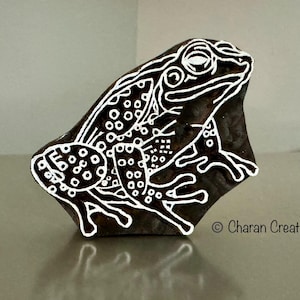 May include: A black wooden block carved with a white outline of a frog. The frog has a detailed design with white dots and lines. The block is a printing stamp.