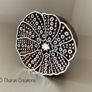 Pode incluir: Um carimbo de impressão de madeira redondo com um desenho floral branco. O desenho apresenta uma estrela central rodeada por um padrão circular de pontos e linhas.